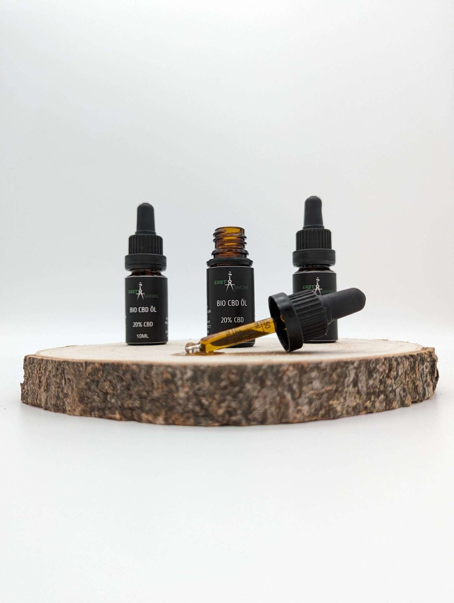 Drei kleine Flaschen Premium Bio CBD Öl mit Pipette stehen auf einer runden Holzplatte, eine Flasche ist geöffnet.