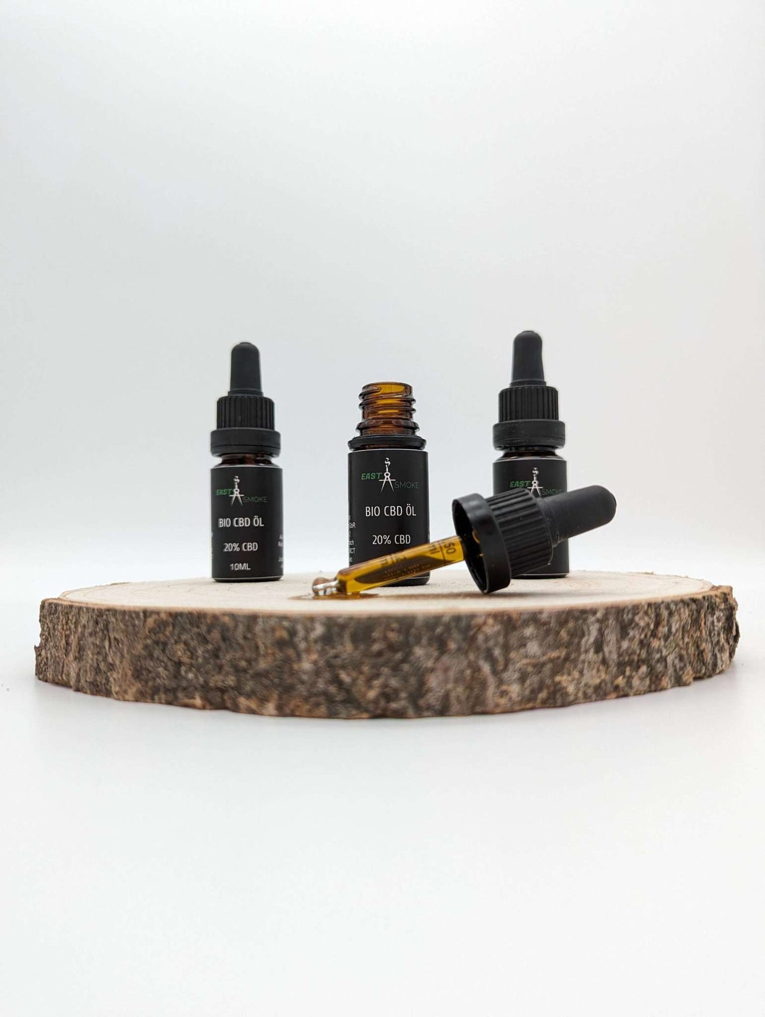 Drei kleine Flaschen Premium Bio CBD Öl mit schwarzem Etikett und Pipetten, eine Flasche offen, auf Holzscheibe platziert.