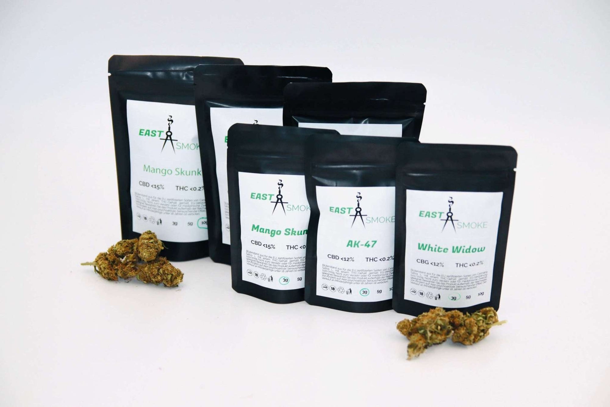 Das Bild zeigt fünf schwarze, aufrecht stehende Verpackungen mit weißen Etiketten, auf denen verschiedene CBD-Blütensorten wie „Mango Skunk“, „AK-47“ und „White Widow“ von der Marke „EASTSMOKE“ zu erkennen sind. Auf den Etiketten stehen Angaben zu dem CBD- und THC-Gehalt. Vor den Verpackungen liegen mehrere grüne bis hellbraune, getrocknete Hanfblüten. Der Hintergrund ist weiß und minimalistisch gehalten, sodass die Produkte im Mittelpunkt stehen.