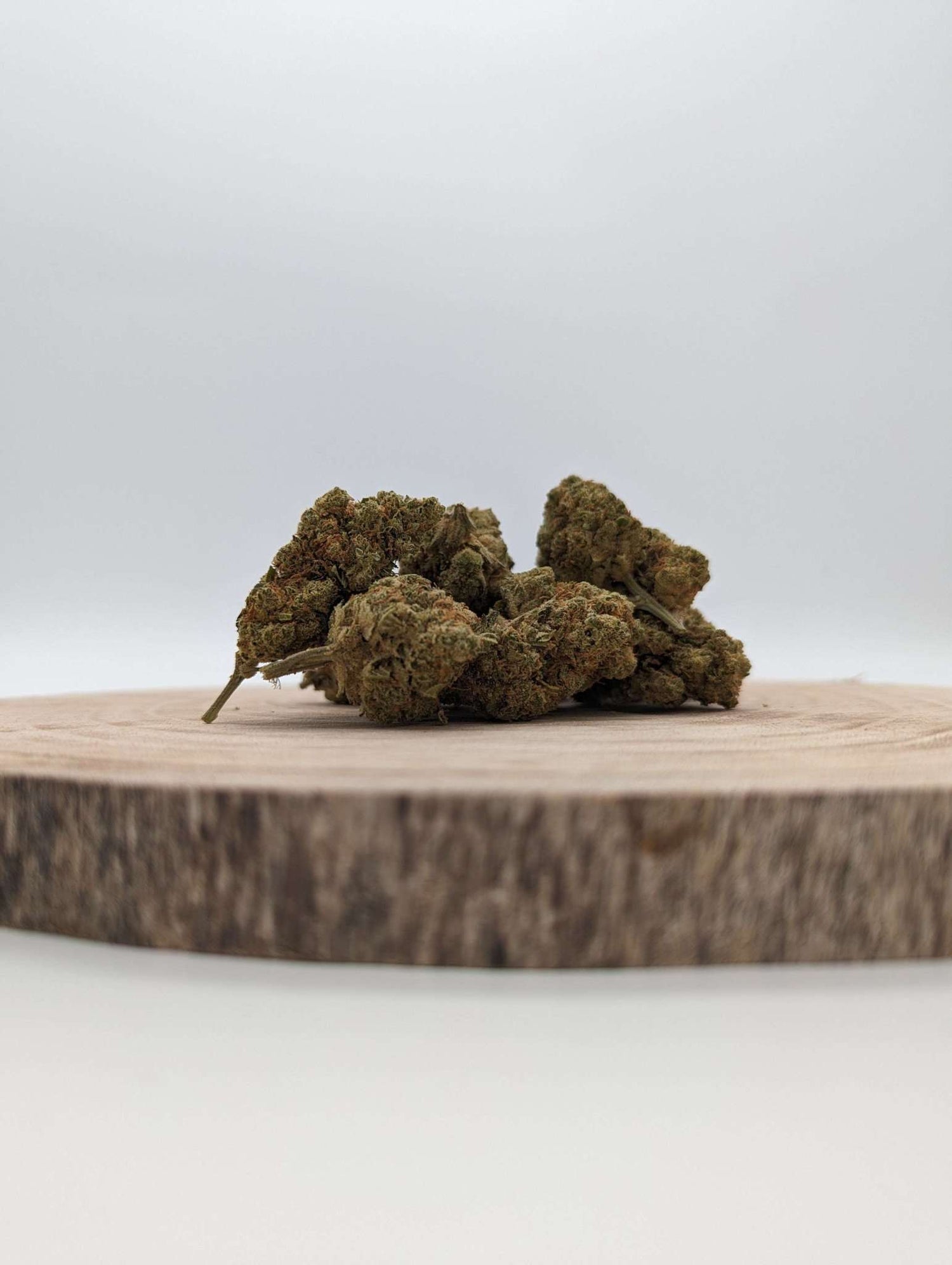 Auf dem Bild sind mehrere grüne, kompakte Pflanzenknospen mit braunen und orangefarbenen Haaren zu sehen. Diese liegen auf einer runden Holzscheibe. Die Oberfläche der Knospen erscheint strukturiert mit kleinen, feinen Kristallen. Der Hintergrund ist neutral und hell, wodurch der Fokus klar auf den Knospen und der Holzscheibe liegt.